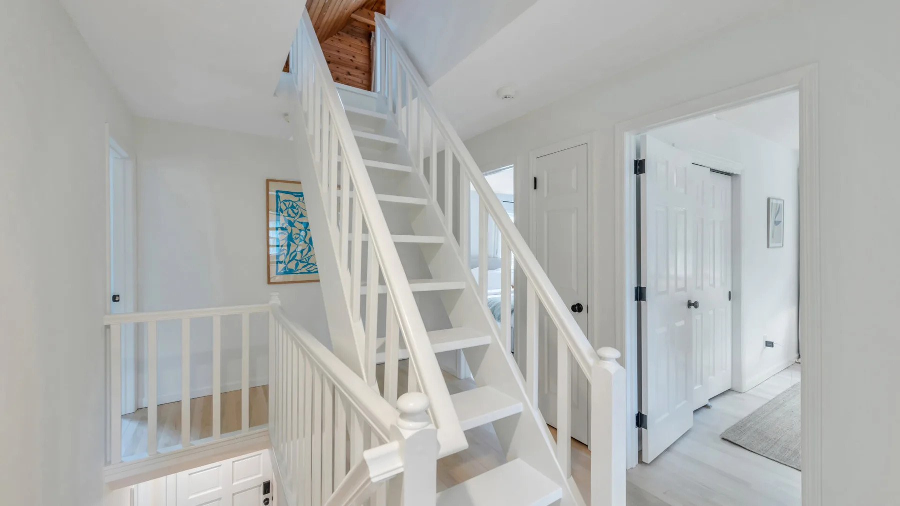 Un pasillo interior luminoso tiene una escalera blanca y un techo de madera encima.
