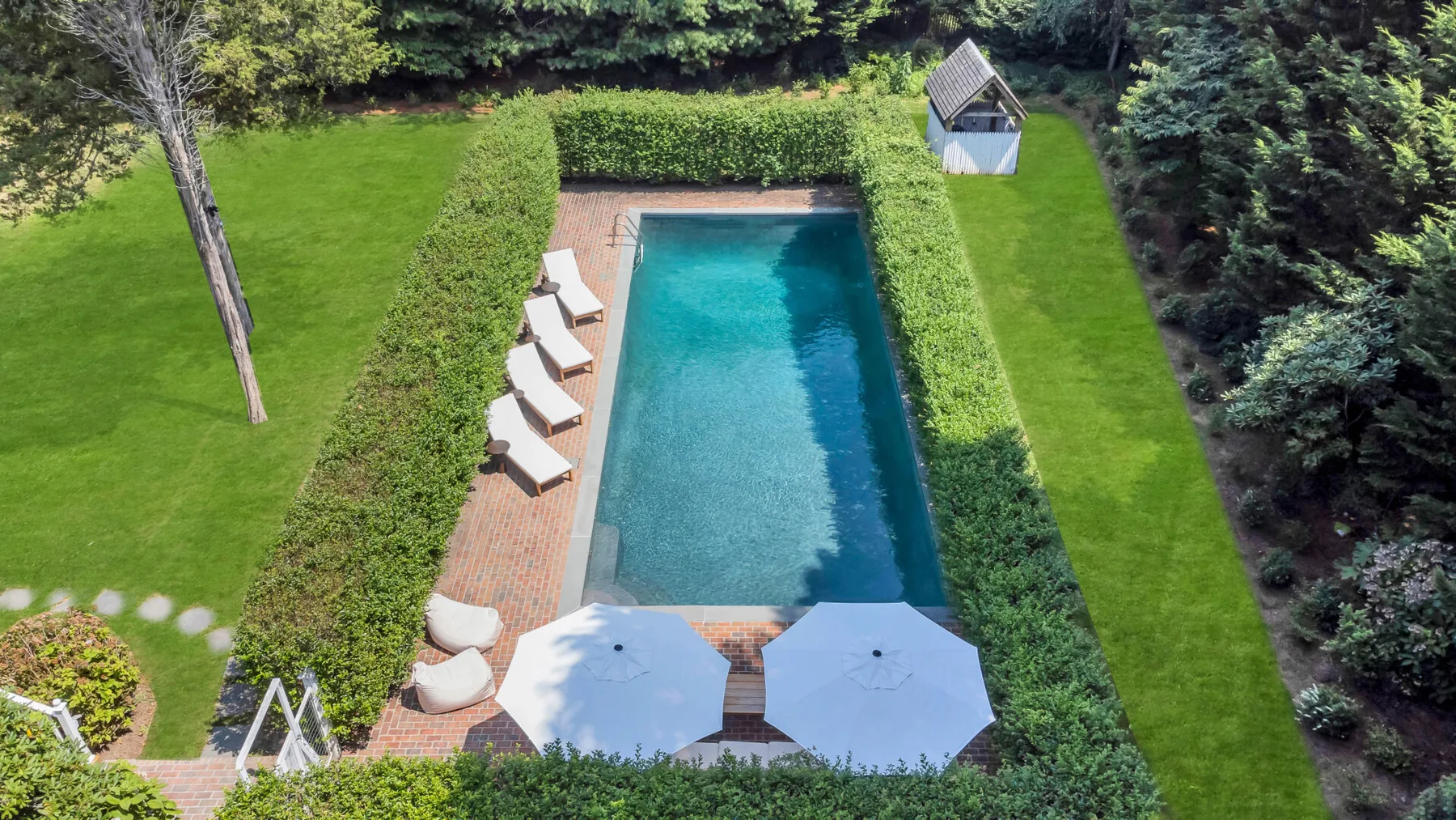 Una piscina rectangular está rodeada de setos verdes, con tumbonas y sombrillas cerca.