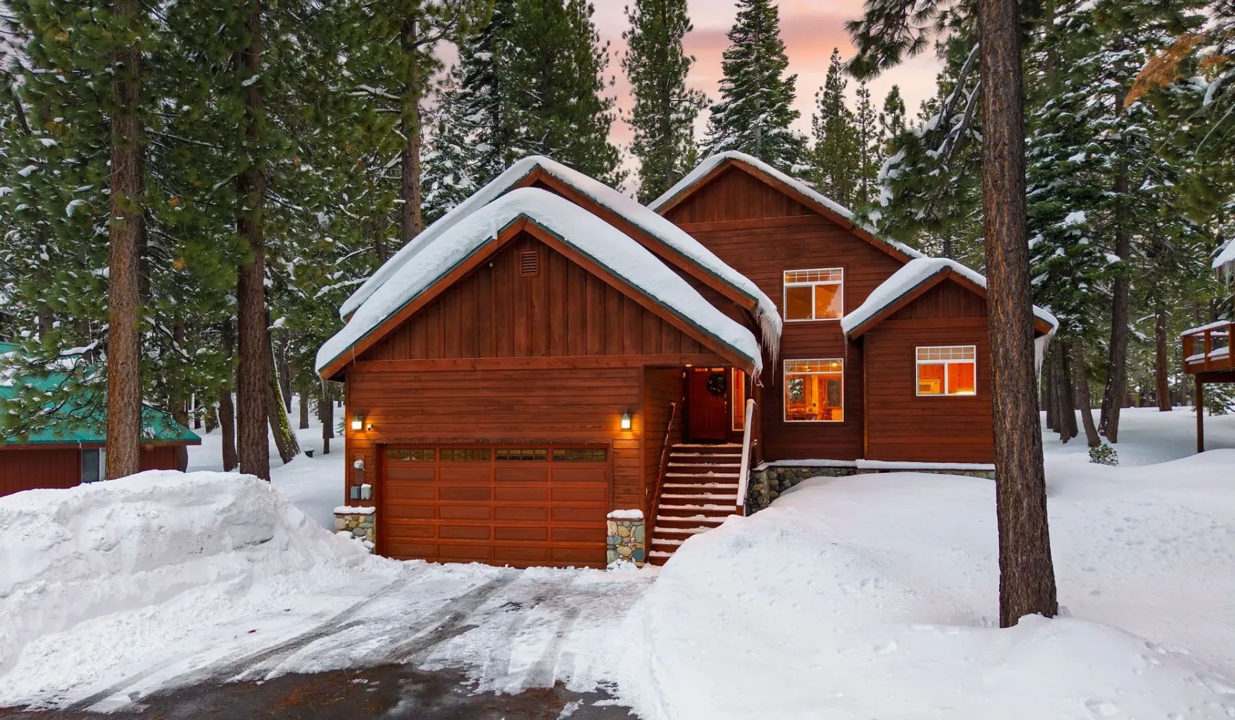 Una cabaña de madera está entre árboles cubiertos de nieve, con nieve acumulada junto a la entrada.