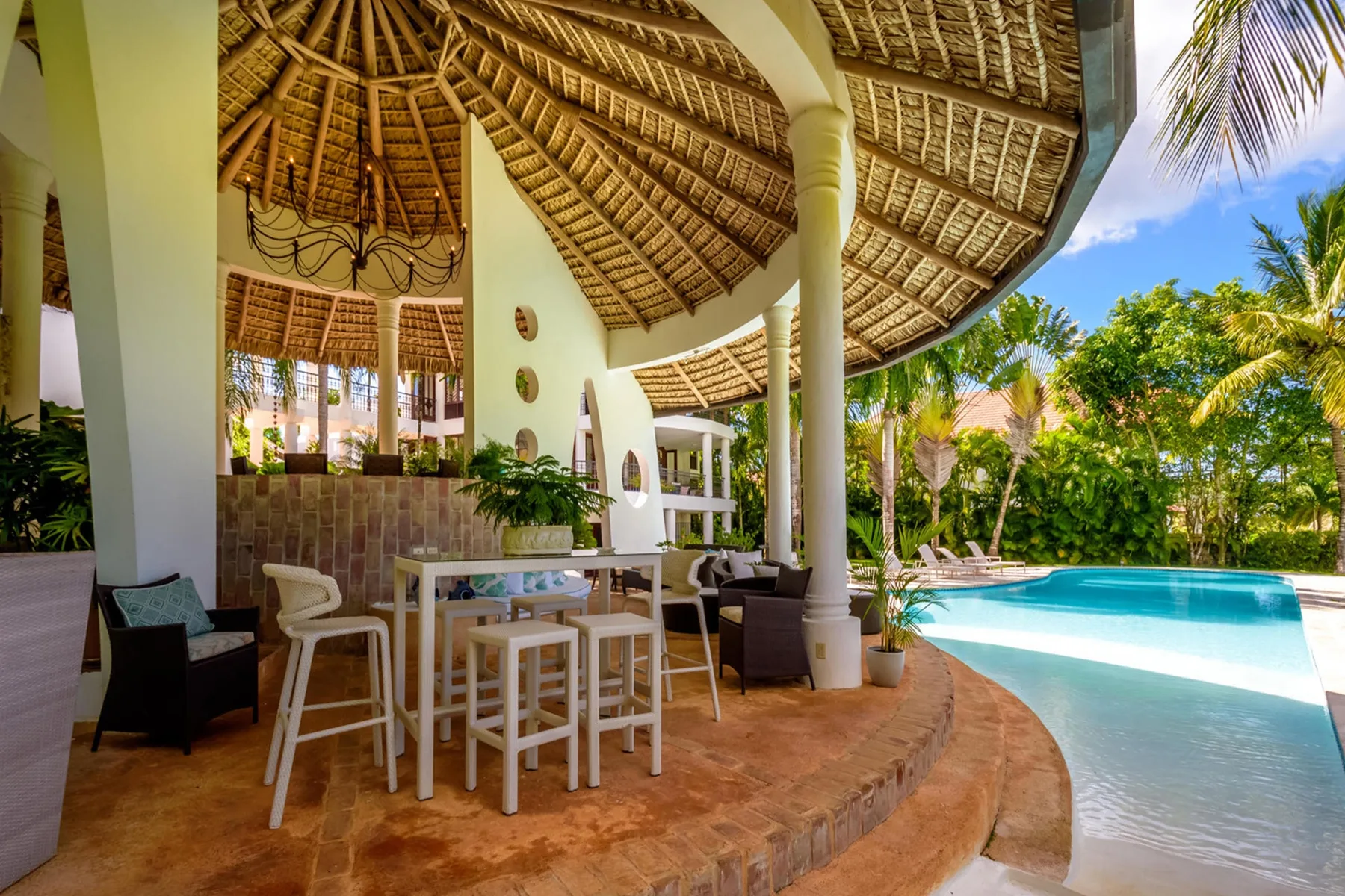 Una zona de comedor al aire libre con techo de paja da a una piscina.