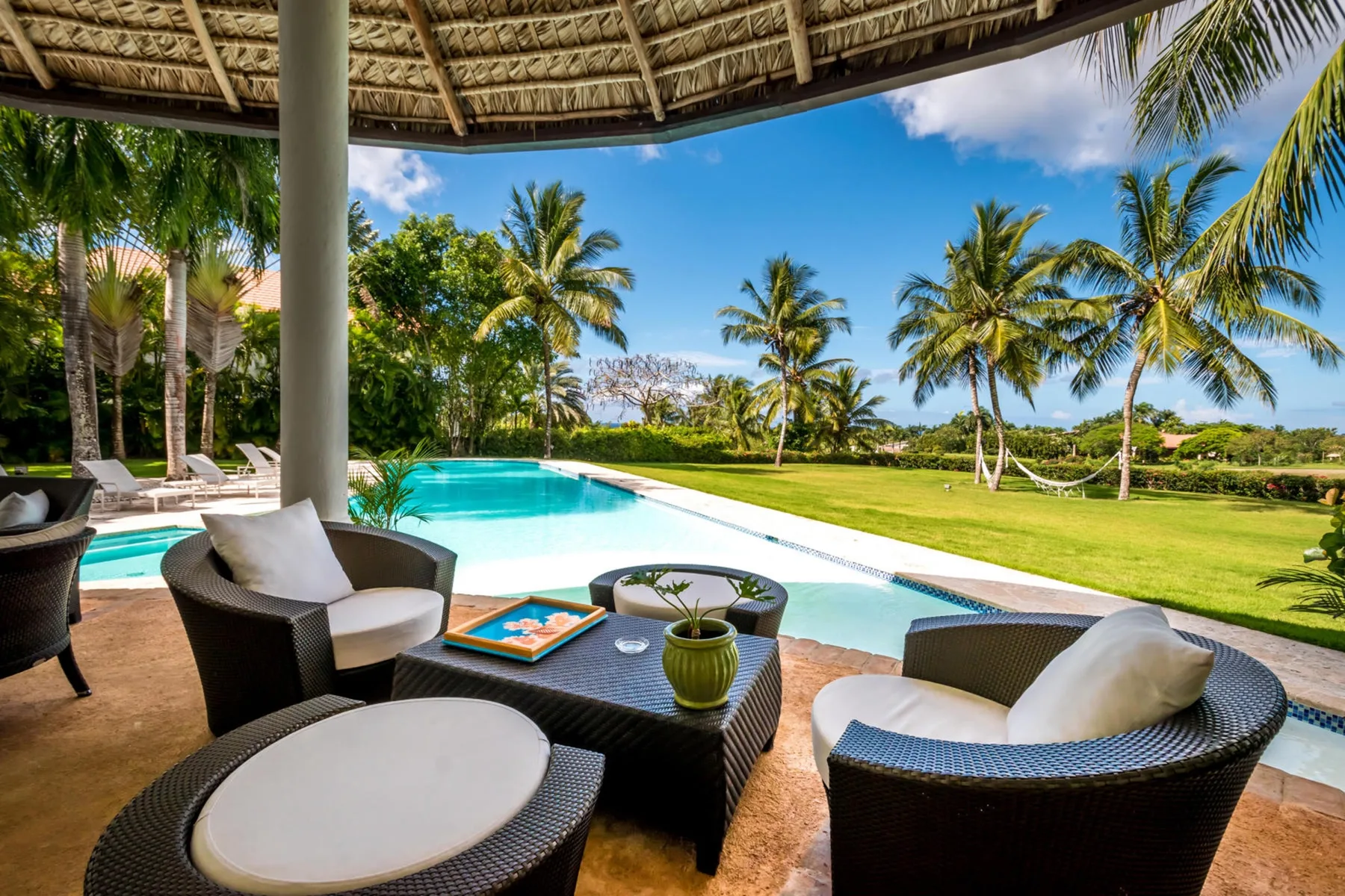 Un patio sombreado con asientos da a una piscina y un césped bordeado de palmeras.