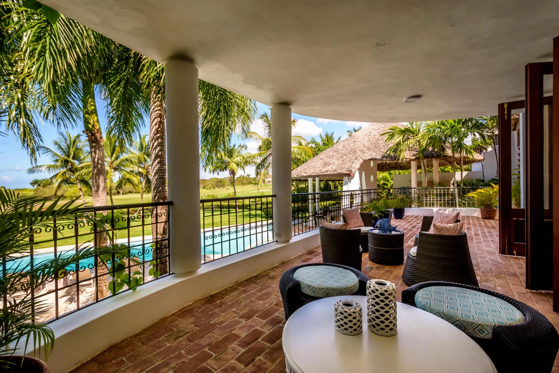 Una terraza cubierta con asientos da a una piscina y palmeras.