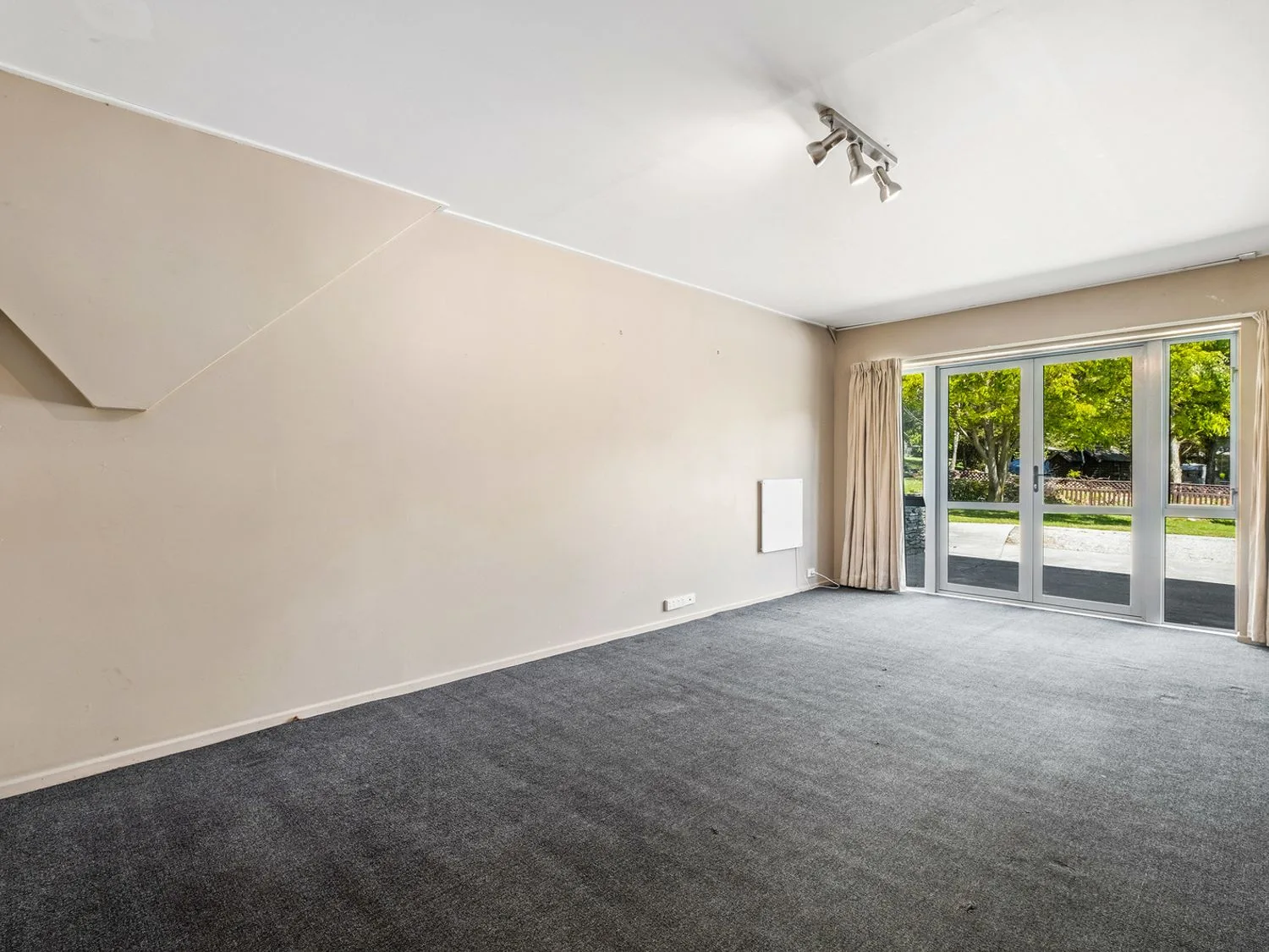 Una habitación vacía con suelo de moqueta gris, paredes beige, un conjunto de puertas de vidrio con cortinas y luces de techo en Taha Wai - Wanaka Holiday Home, en Wanaka.