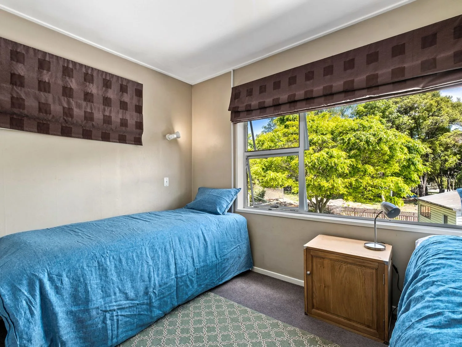 Un dormitorio con dos camas individuales, colchas azules, una mesa de noche de madera y una ventana con vistas a árboles verdes en Taha Wai - Wanaka Holiday Home, en Wanaka.