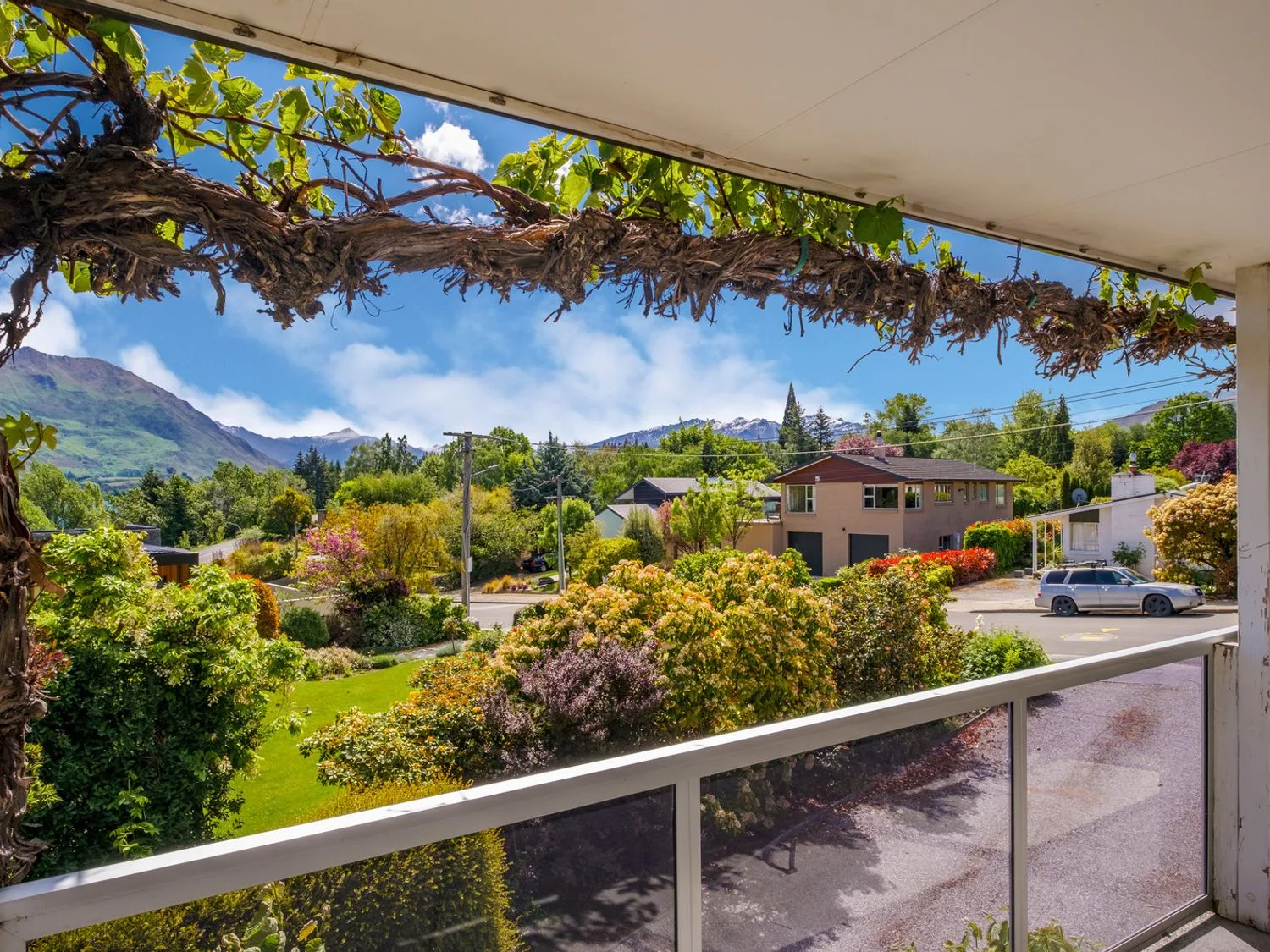 Un porche da a un jardín cuidado, casas cercanas y montañas bajo un cielo despejado.