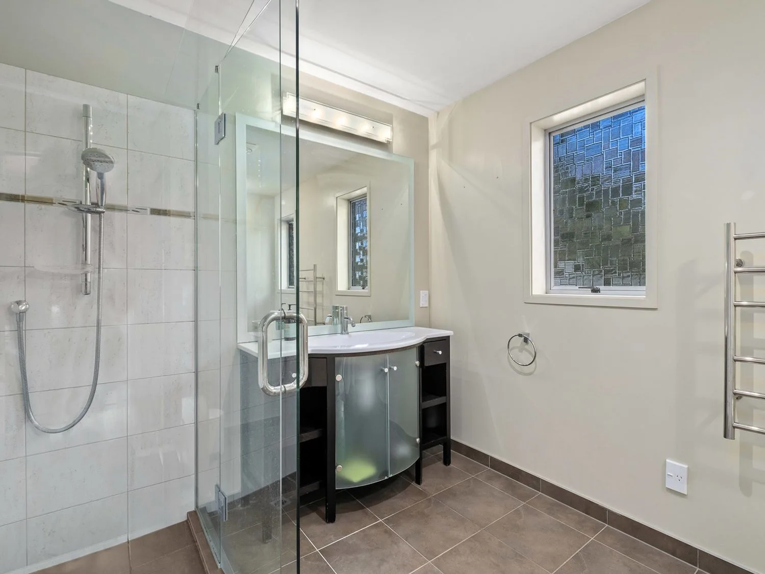 Un baño con una ducha con mampara de vidrio, un mueble de lavabo con espejo y puertas de vidrio esmerilado, una ventana y un toallero en Taha Wai - Wanaka Holiday Home, en Wanaka.