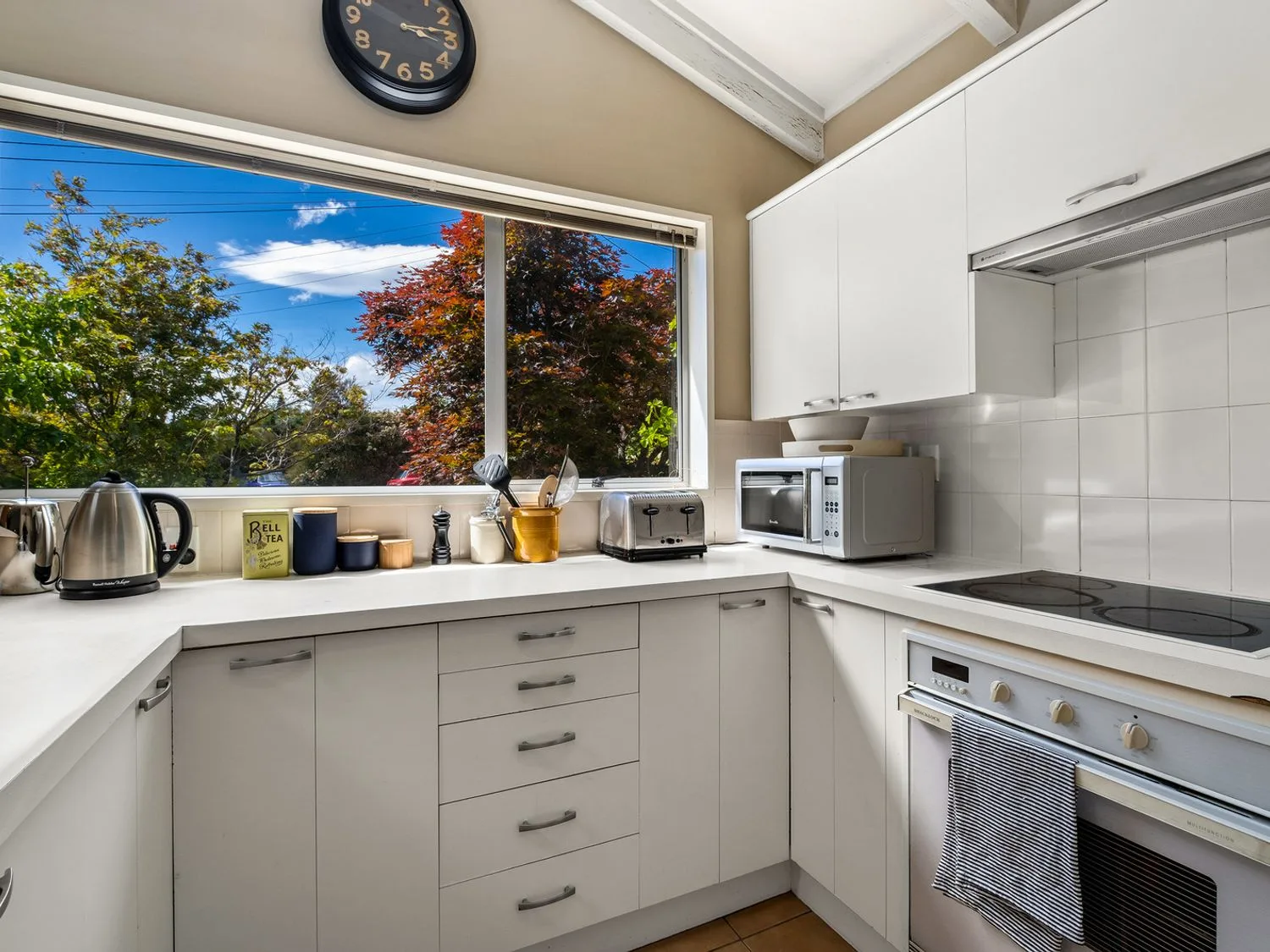 Una cocina con armarios blancos, encimera, microondas, tostadora, hervidor y utensilios cerca de una ventana con árboles en el exterior en Taha Wai - Wanaka Holiday Home, en Wanaka.
