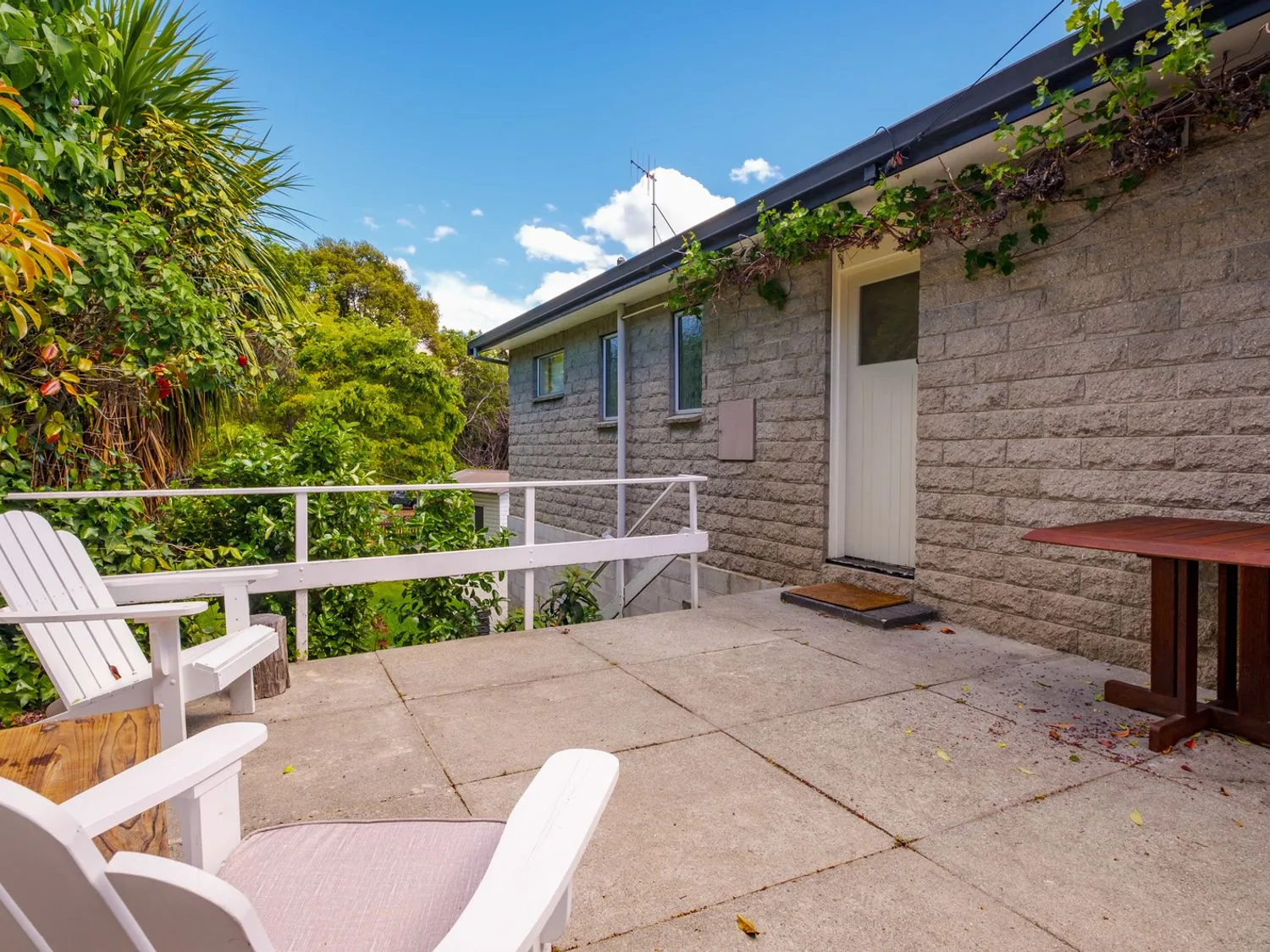 Un patio de hormigón con sillas blancas y una mesa de madera junto a una casa de piedra con una puerta blanca y plantas verdes alrededor en Taha Wai - Wanaka Holiday Home, en Wanaka.