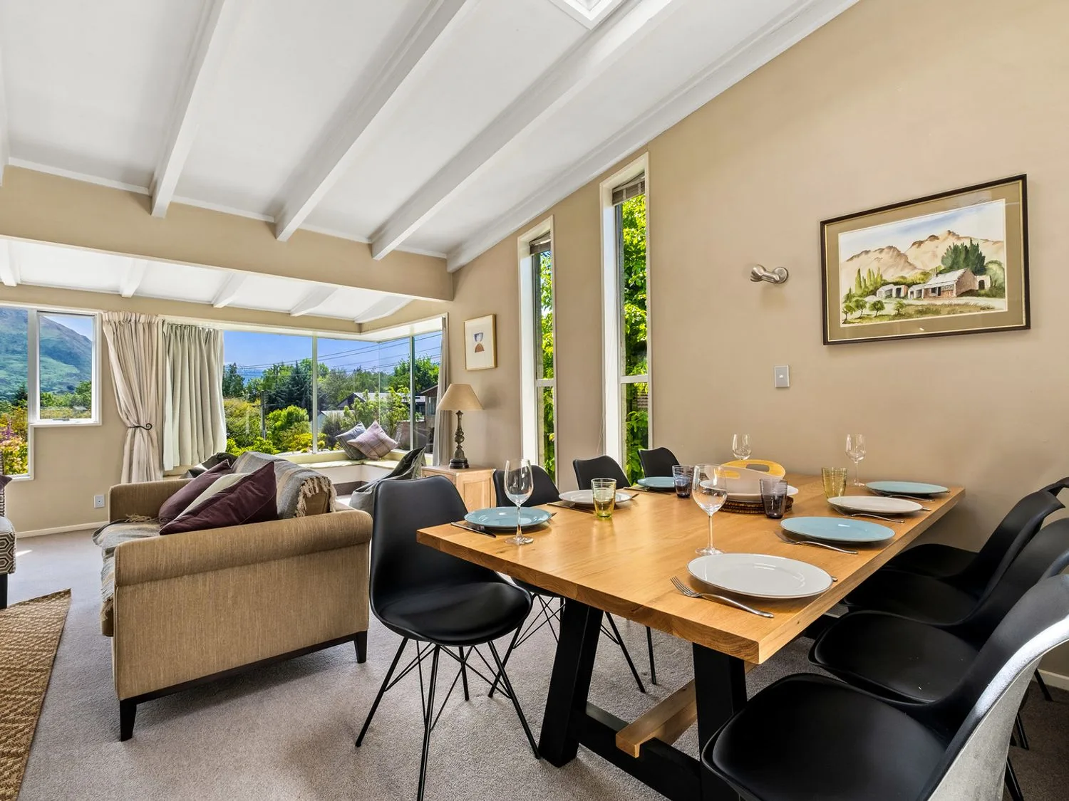 Una zona de comedor con una mesa de madera puesta con platos y vasos, junto a una sala de estar con sofás cerca de grandes ventanas en Taha Wai - Wanaka Holiday Home, en Wanaka.