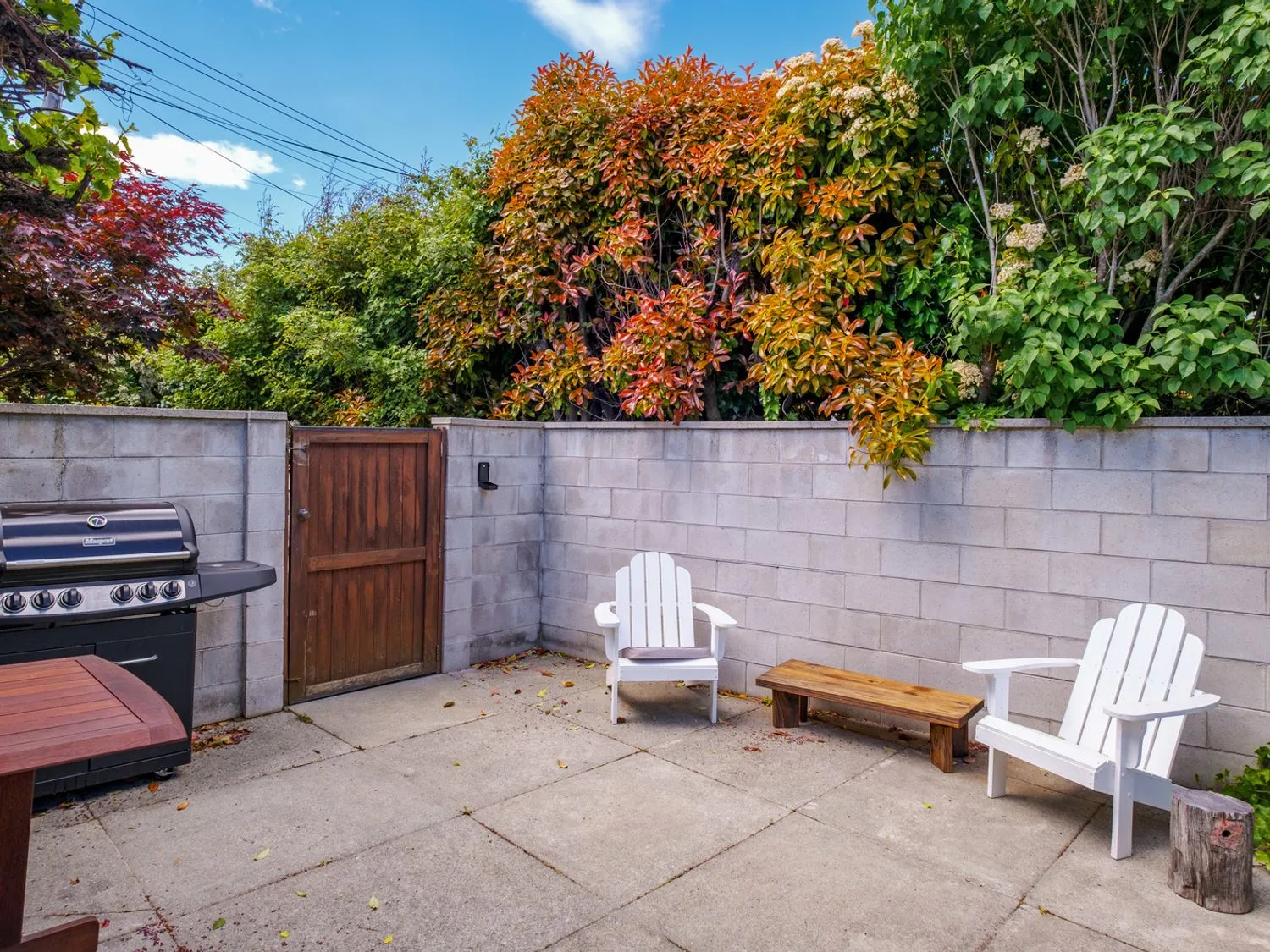 Una zona de patio exterior con dos sillas blancas, un banco de madera, una parrilla y una puerta de madera, rodeada por un muro de bloques de hormigón y vegetación en Taha Wai - Wanaka Holiday Home, en Wanaka.
