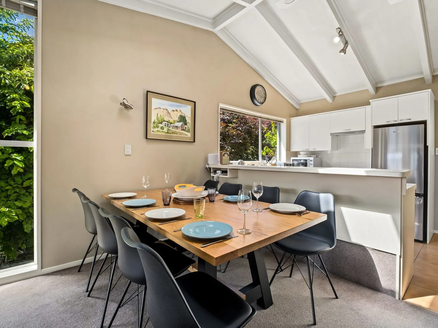 Una zona de comedor con una mesa de madera puesta con platos y vasos y sillas negras, junto a una cocina con armarios blancos y un refrigerador de acero inoxidable en Taha Wai - Wanaka Holiday Home, en Wanaka.
