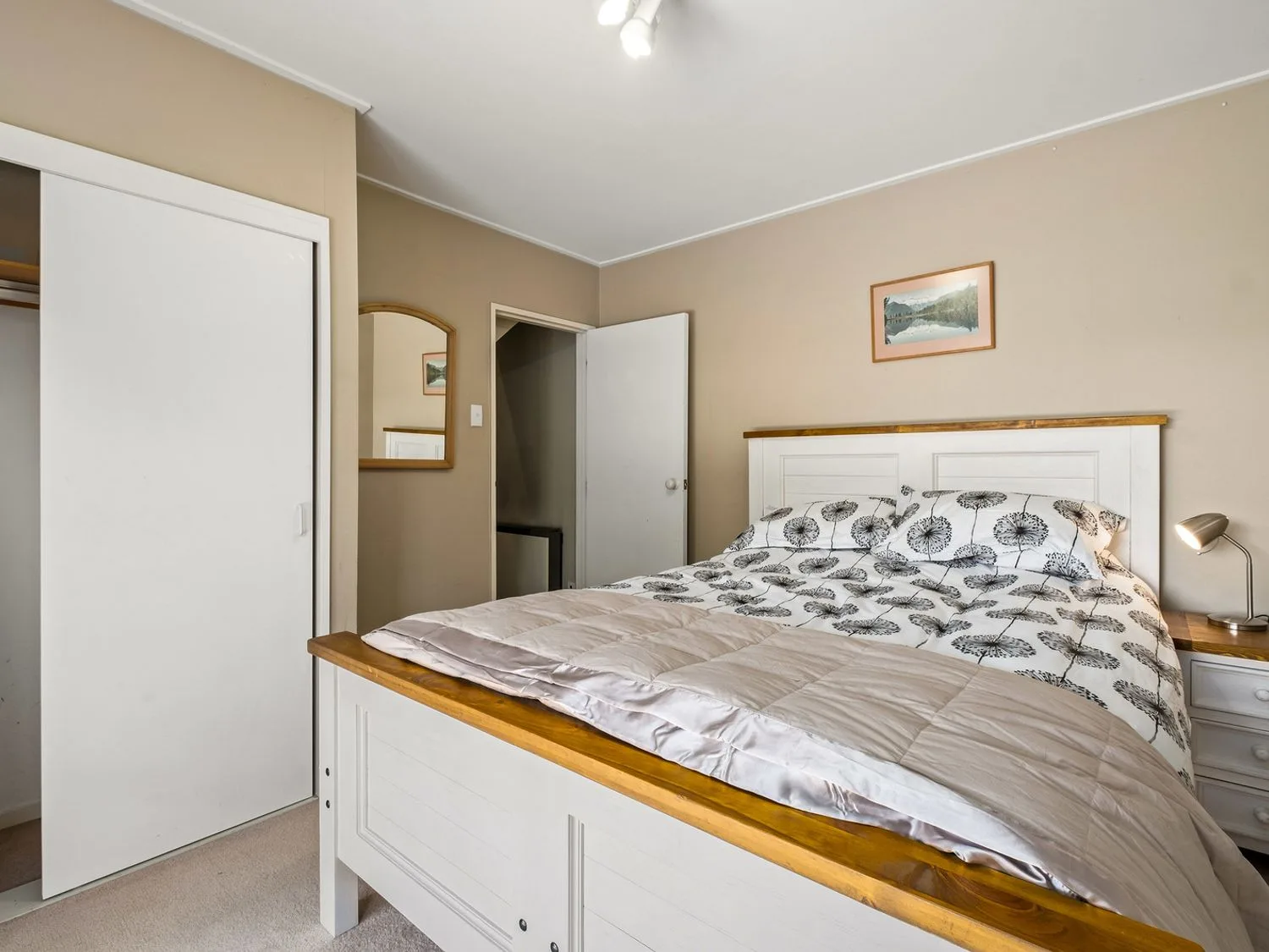 Un dormitorio con una cama blanca y de madera, un edredón estampado, una mesa de noche con una lámpara, un espejo y una puerta abierta en Taha Wai - Wanaka Holiday Home, en Wanaka.