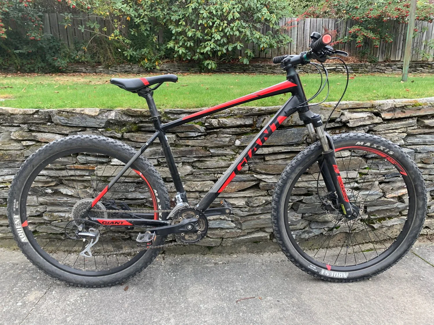 Una bicicleta de montaña negra y roja se apoya contra un muro bajo de piedra junto a un jardín con césped.