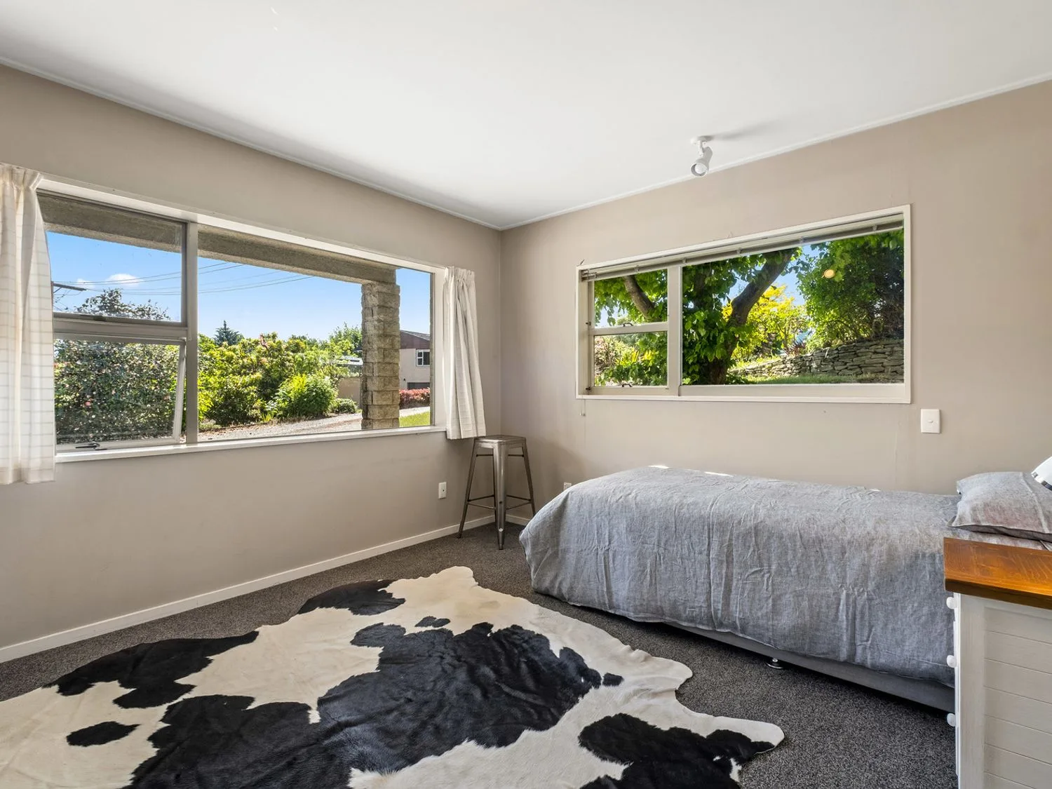 Un dormitorio con una cama individual, una alfombra de piel de vaca, un taburete de metal y dos grandes ventanas con vistas al jardín en Taha Wai - Wanaka Holiday Home, en Wanaka.