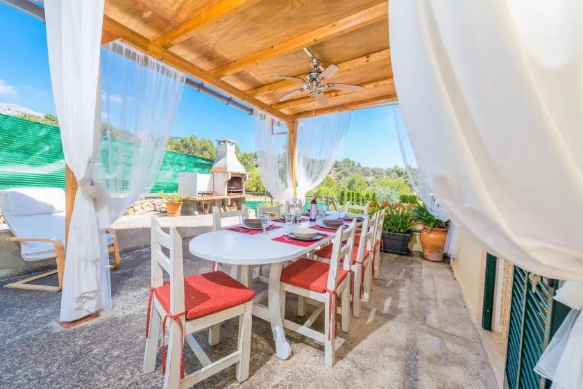 A covered outdoor dining area has a long table and white chairs with red cushions.