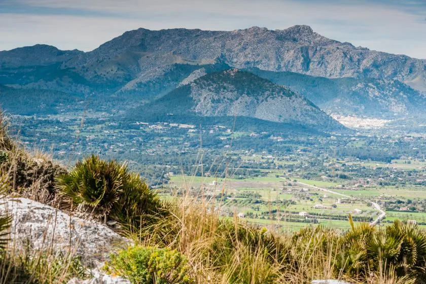 A mountain range rises above a broad valley with fields and scattered buildings.