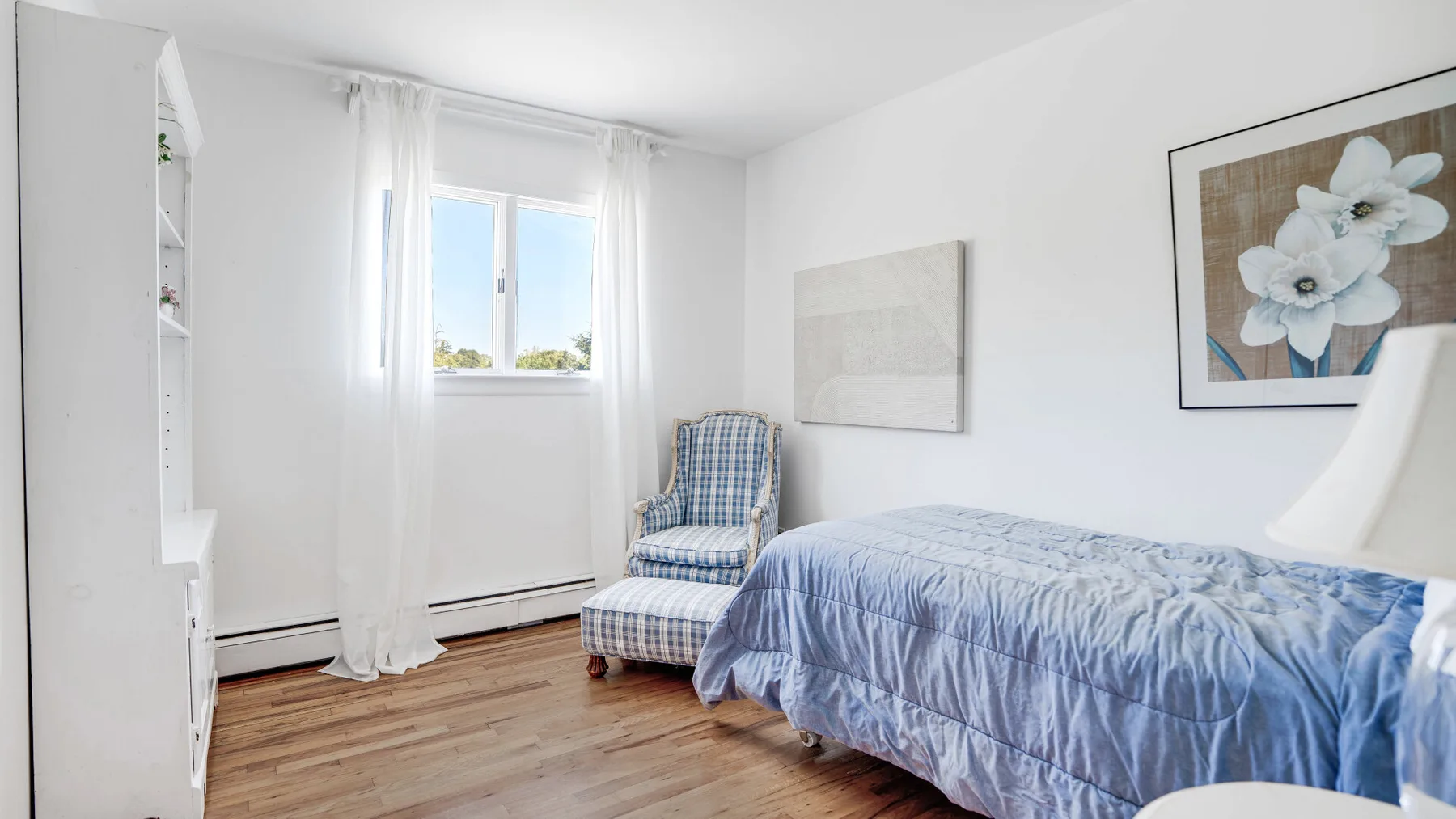 Un dormitorio luminoso con una cama cubierta de azul y una ventana con cortinas translúcidas.