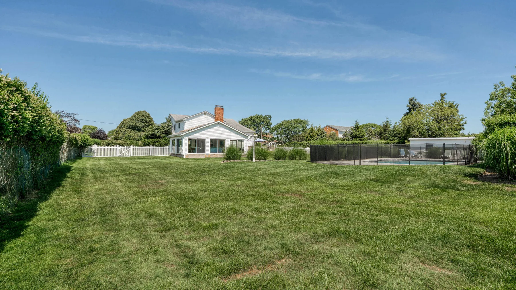 A broad grassy backyard stretches toward a house, with hedges and a fenced area.