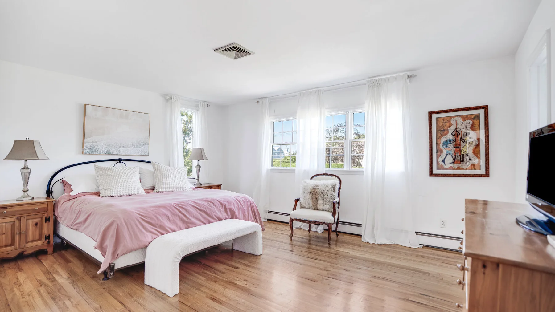 Un dormitorio luminoso con una cama cubierta de ropa de cama rosa y ventanas con cortinas translúcidas.