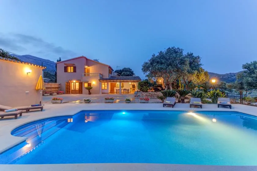 An outdoor swimming pool glows at dusk beside a lit building and landscaped seating area.