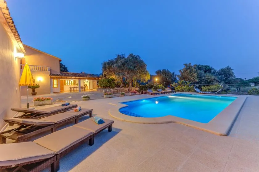 An outdoor swimming pool sits beside a paved patio and lounge seating at dusk.