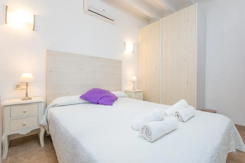 A bright bedroom with a white bed, purple cushion, and pale wood wardrobe.