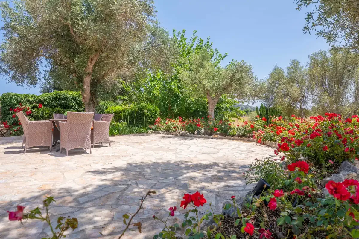 A stone patio with outdoor seating is surrounded by trees and flowering shrubs.