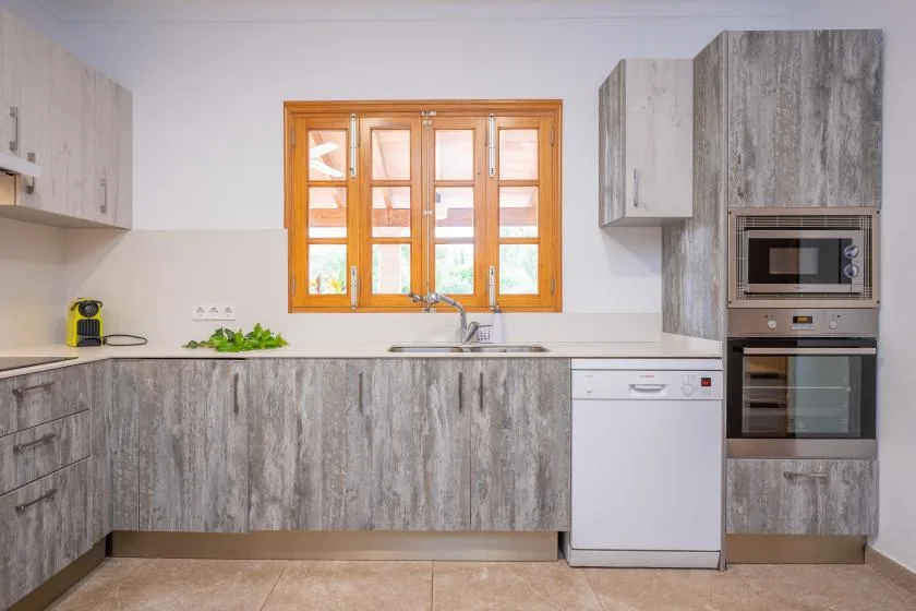 A kitchen with gray cabinets, a sink beneath a wooden-framed window, and built-in appliances.