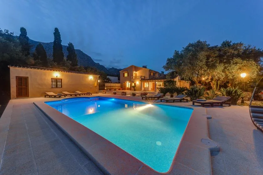 An outdoor swimming pool glows beside a warmly lit building at dusk.