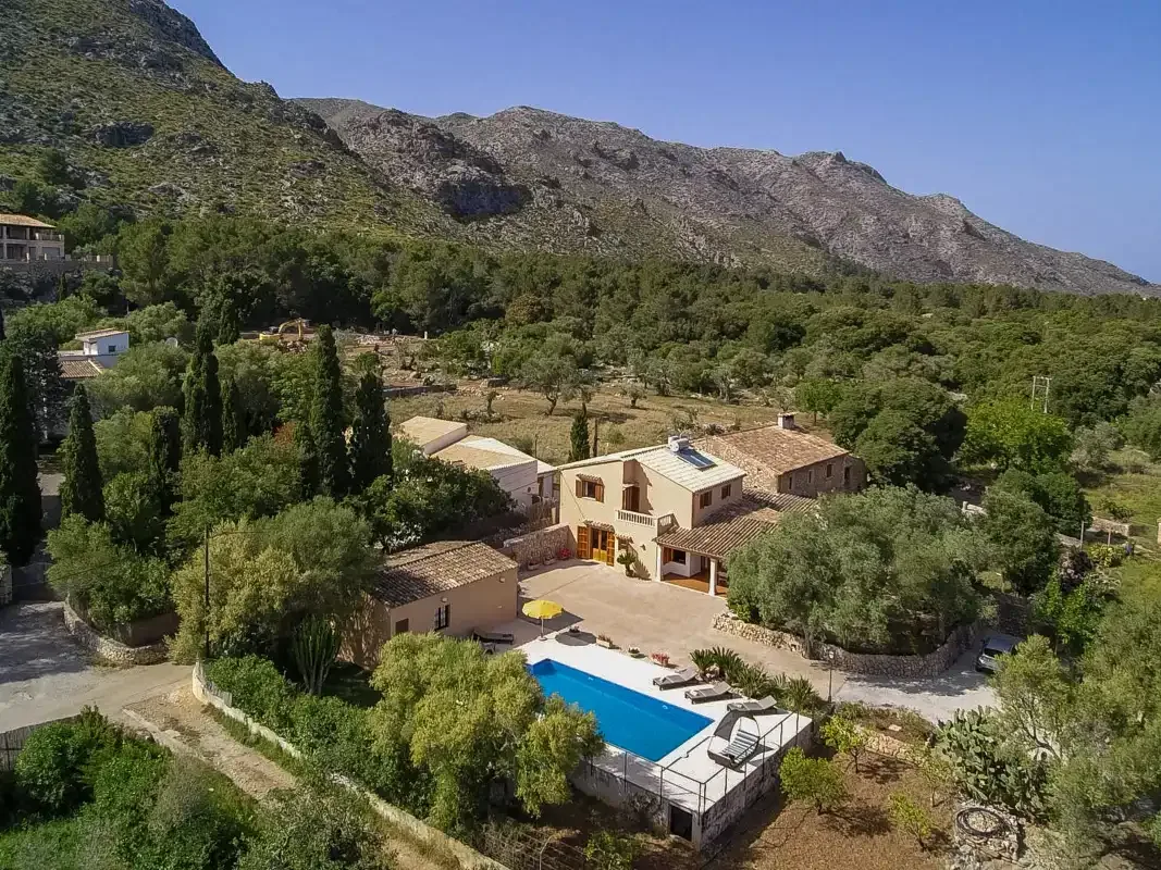 A countryside property with a swimming pool sits among trees beneath rocky hills.