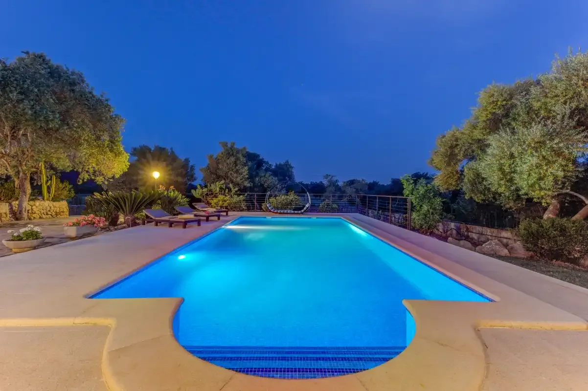 A lit swimming pool sits on a paved patio surrounded by trees.