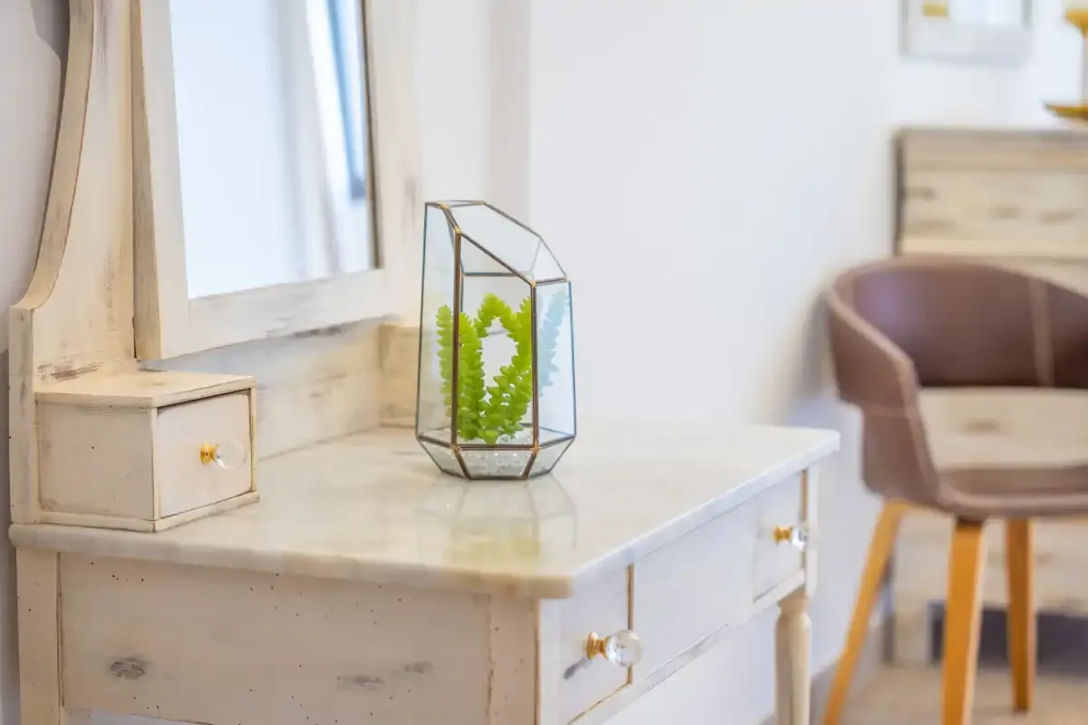 A pale wooden desk with a glass plant terrarium sits beside a window.