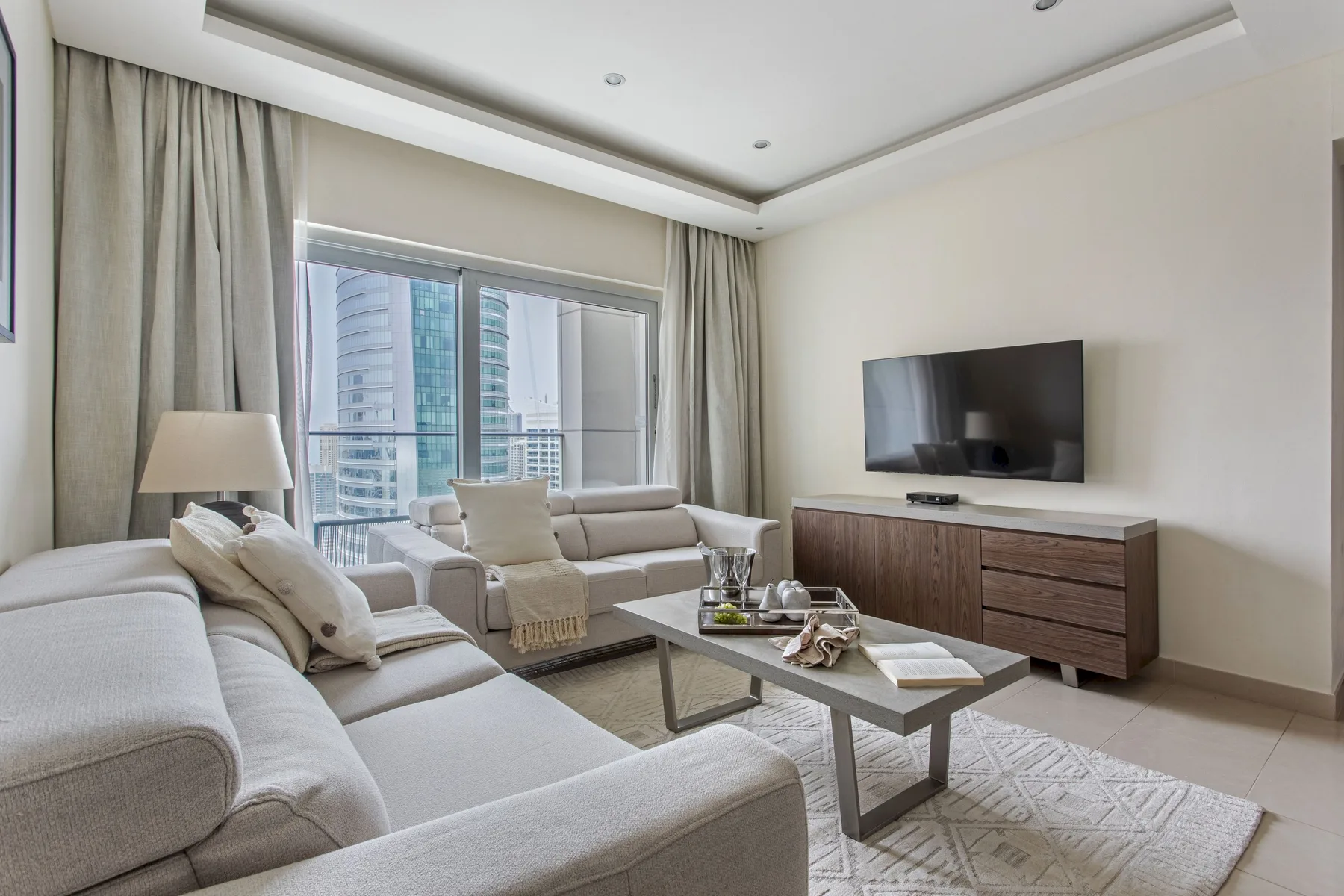Una sala luminosa con asientos, un televisor y grandes ventanales con cortinas.