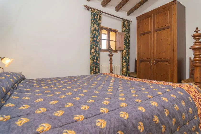 A bedroom with a patterned bedspread, wooden wardrobe, and window with curtains.