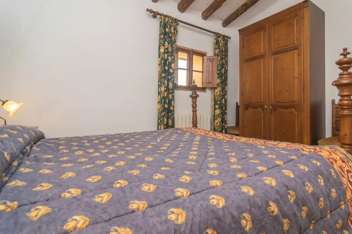 A bedroom with a patterned bedspread, wooden wardrobe, and curtained window.
