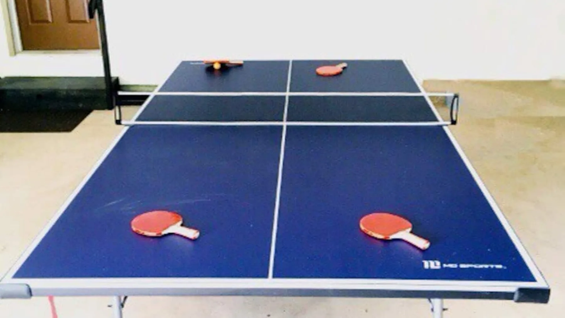 A blue table tennis table with paddles stands in a simple indoor room.