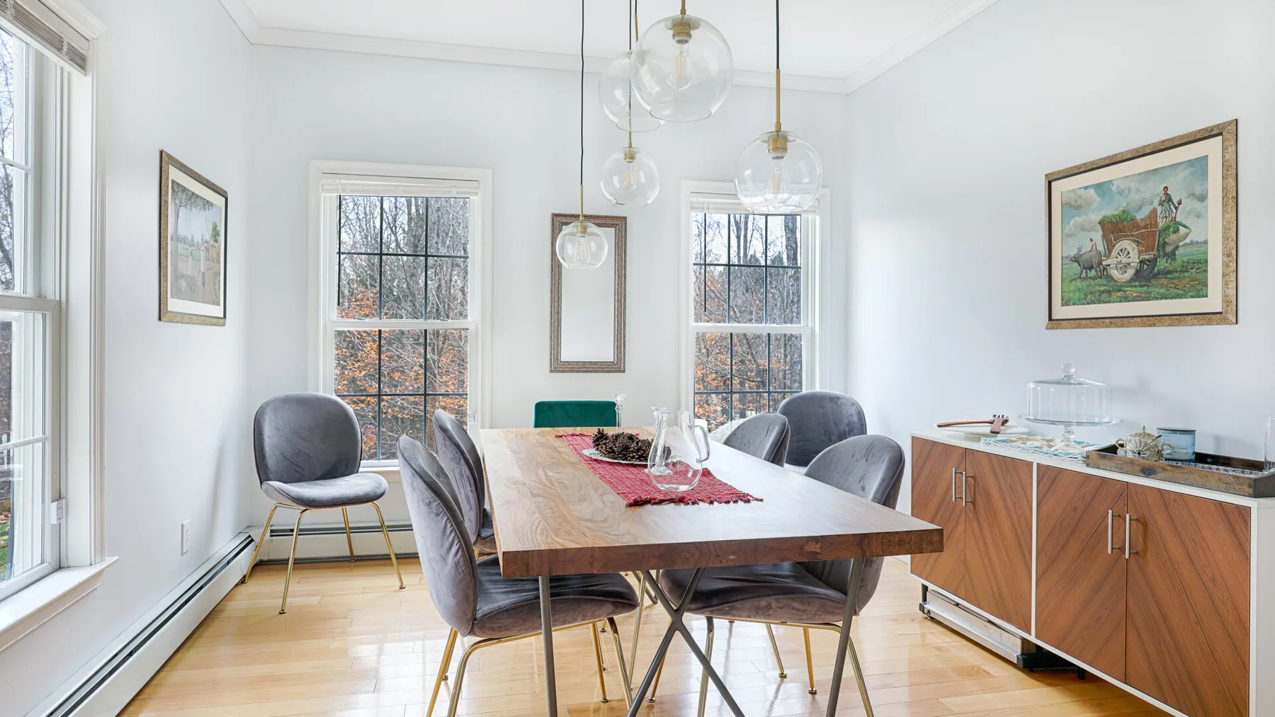 A bright dining room features a long wooden table beneath glass pendant lights.
