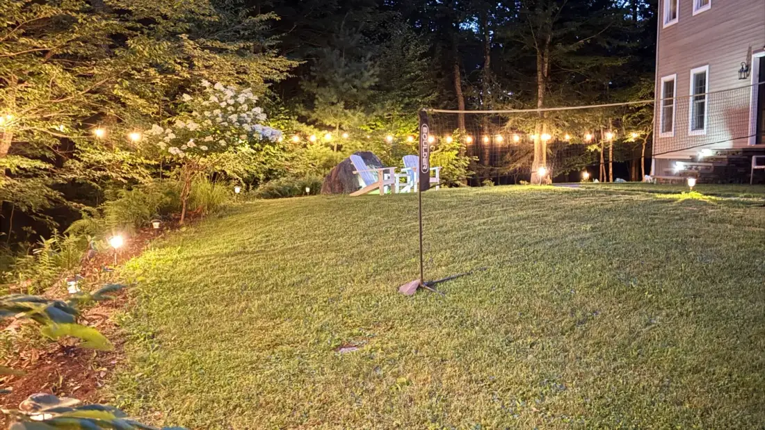 A grassy backyard at night is lit by string lights beside a house.