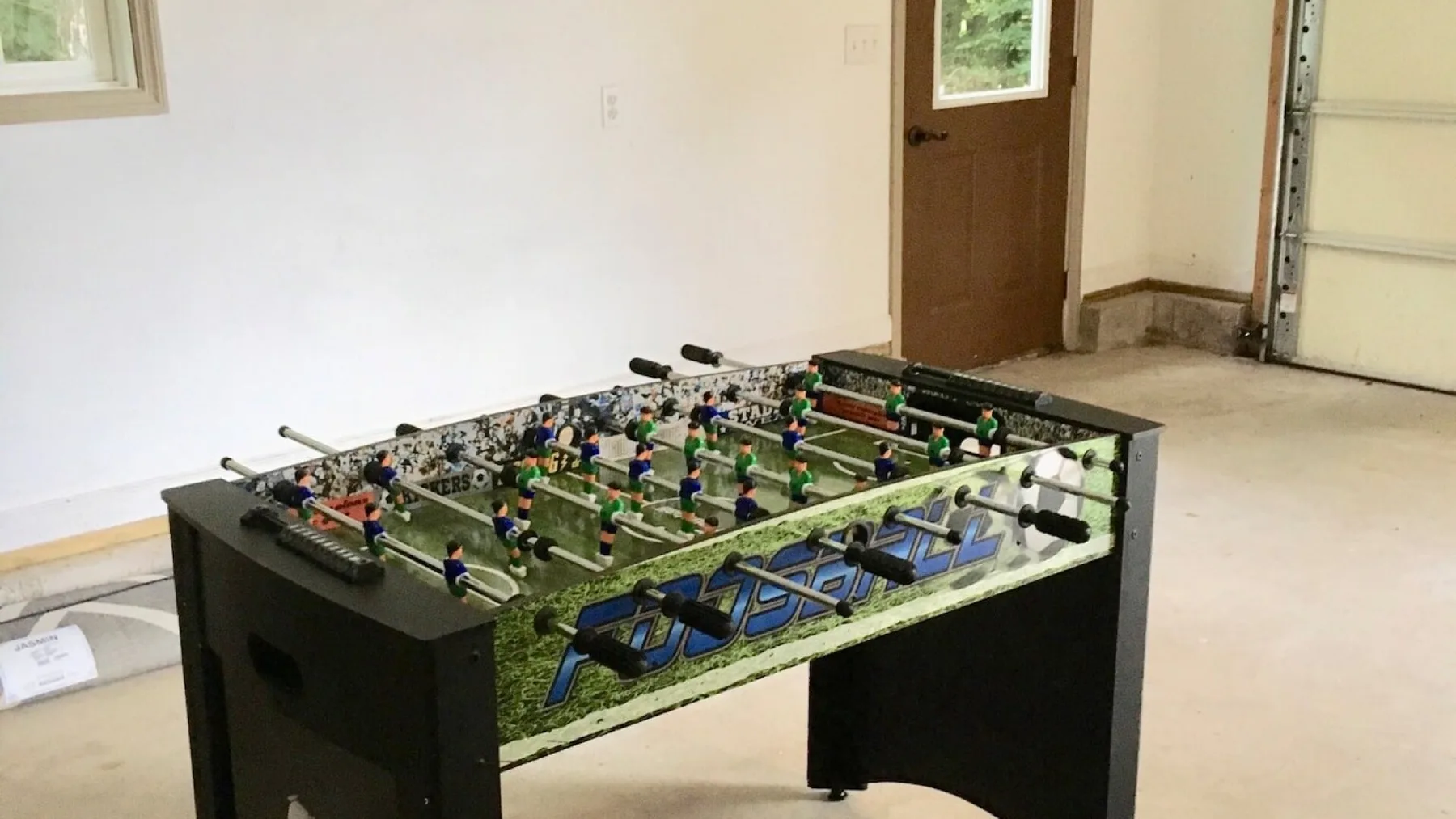 A foosball table stands in a bare garage beside a door.
