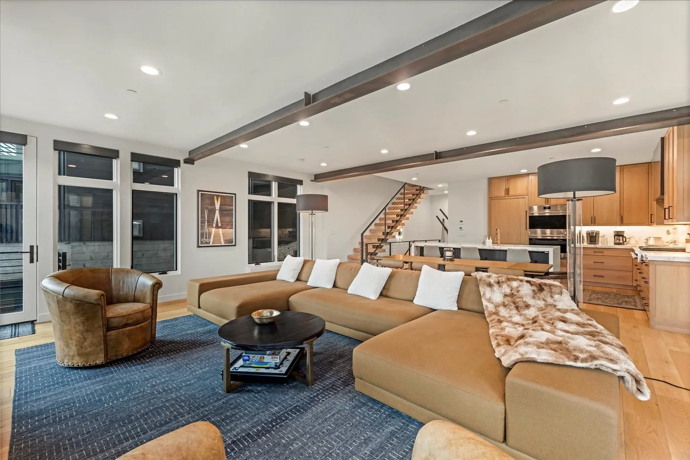 An open-plan living room with sectional seating, a blue rug, and a kitchen beyond.