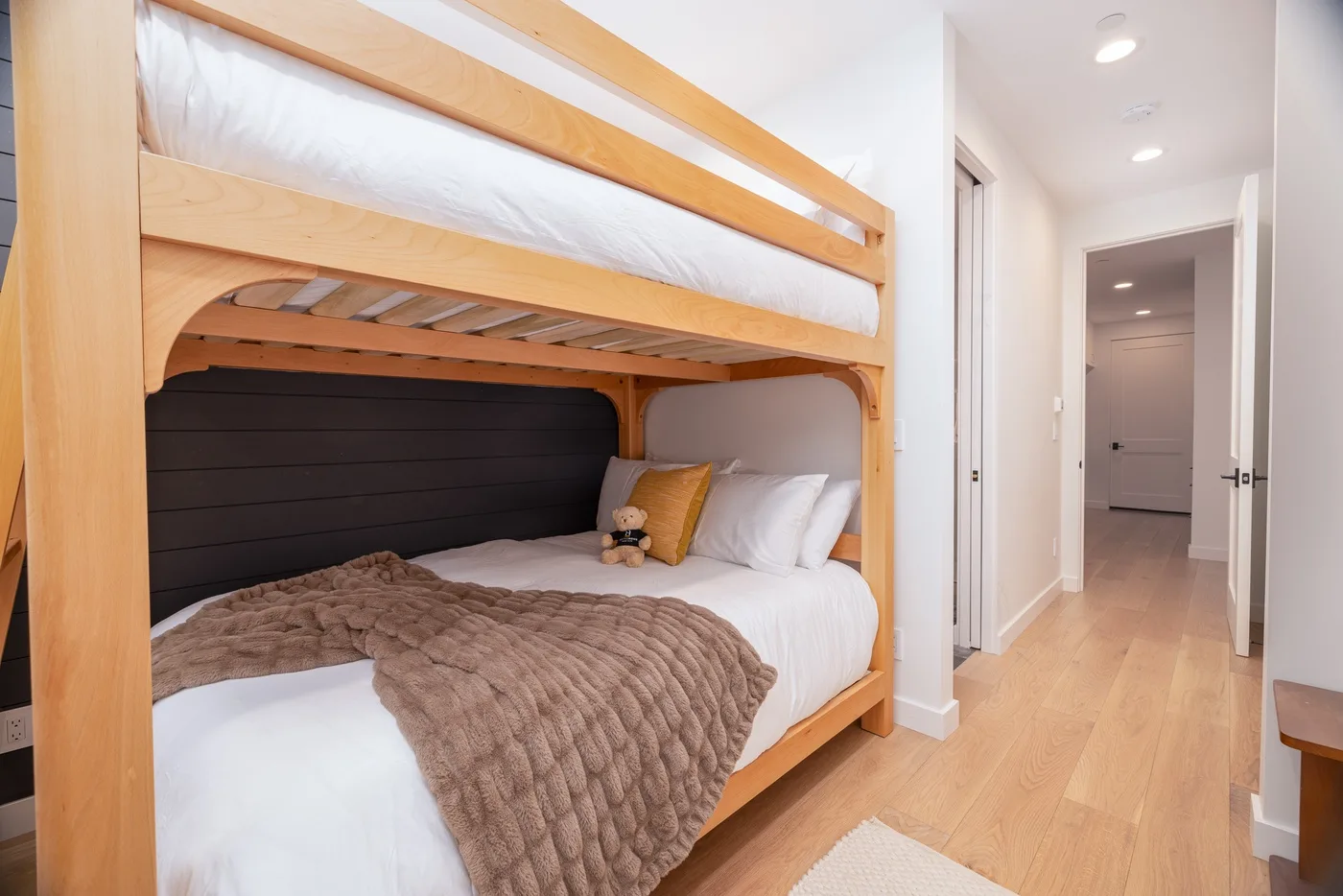 A bedroom with a wooden bunk bed, white bedding, and a hallway beside it.
