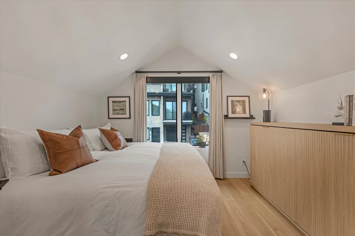 A bedroom with a bed beside a large window and light wood flooring.