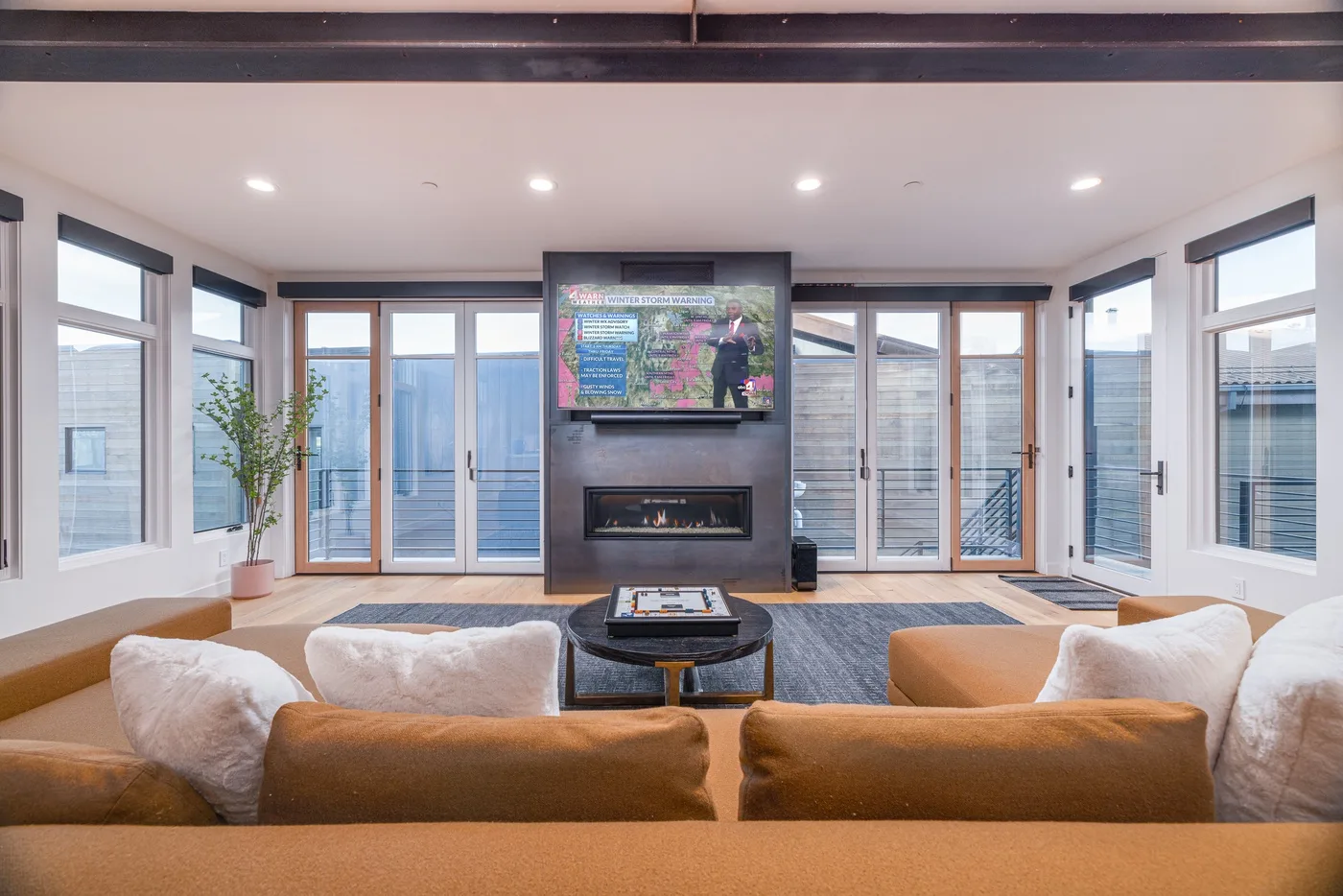 A bright living room has a television and fireplace between large windows.