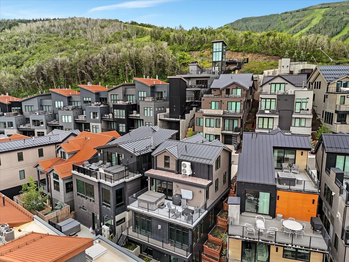 A cluster of modern hillside buildings with balconies sits below a green, wooded ridge.