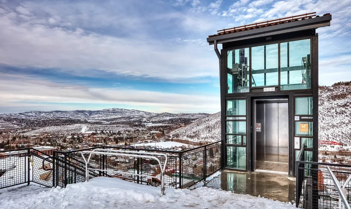 An outdoor observation deck overlooks a snow-covered town through a glass-sided elevator tower.