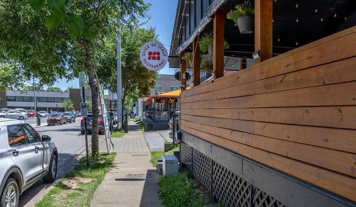Una acera soleada bordea una terraza de comedor de madera en una calle arbolada.
