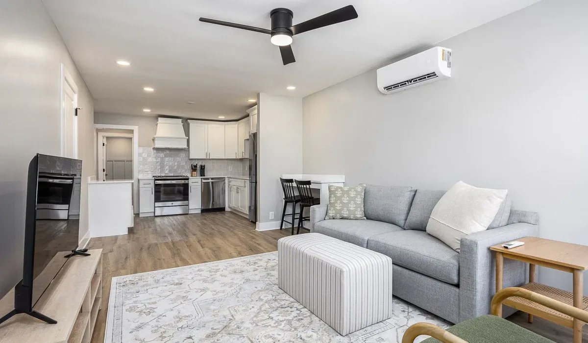 Una sala luminosa se conecta con una cocina compacta, con un sofá gris y un televisor.