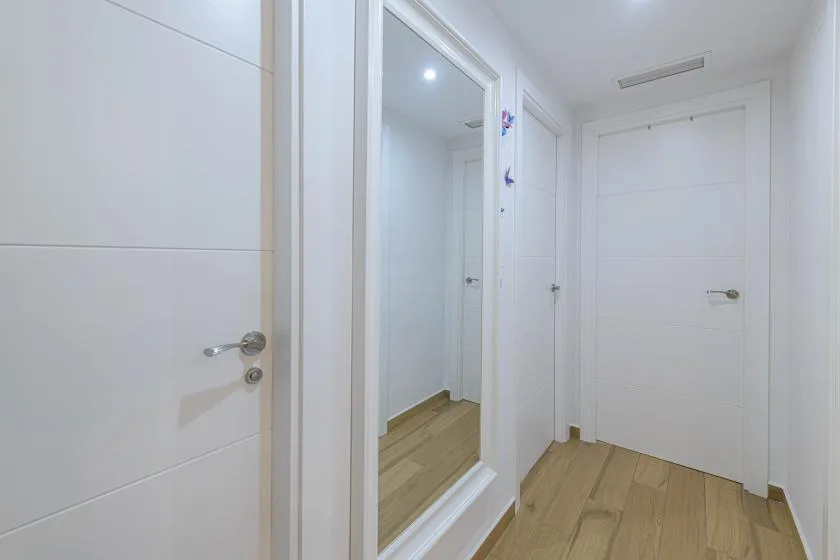 Un pasillo luminoso con puertas blancas, un espejo de cuerpo entero y suelo de madera.