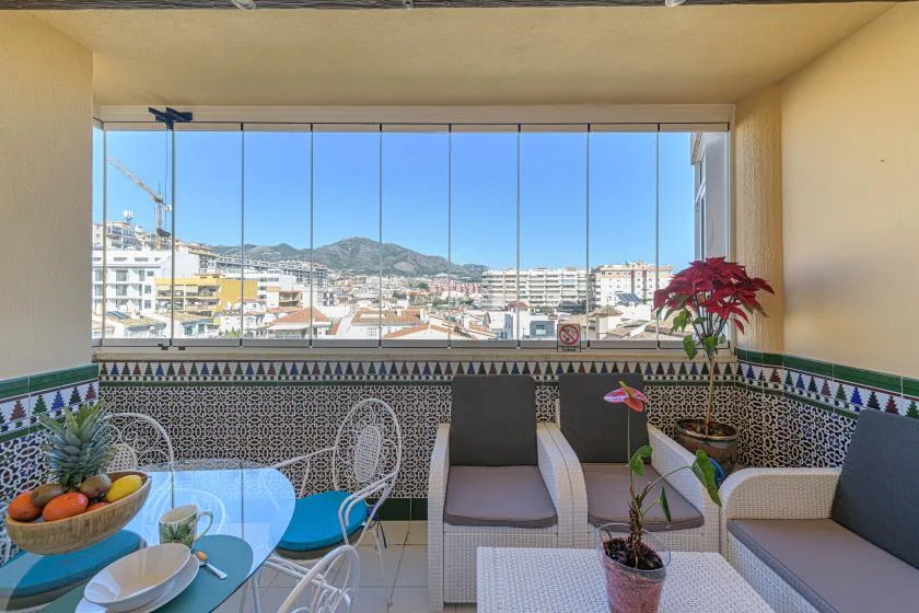 A bright enclosed balcony with seating, patterned tile, and a city view.