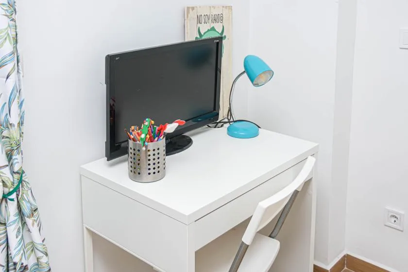 Un escritorio blanco con un monitor, bolígrafos de colores y una lámpara azul.